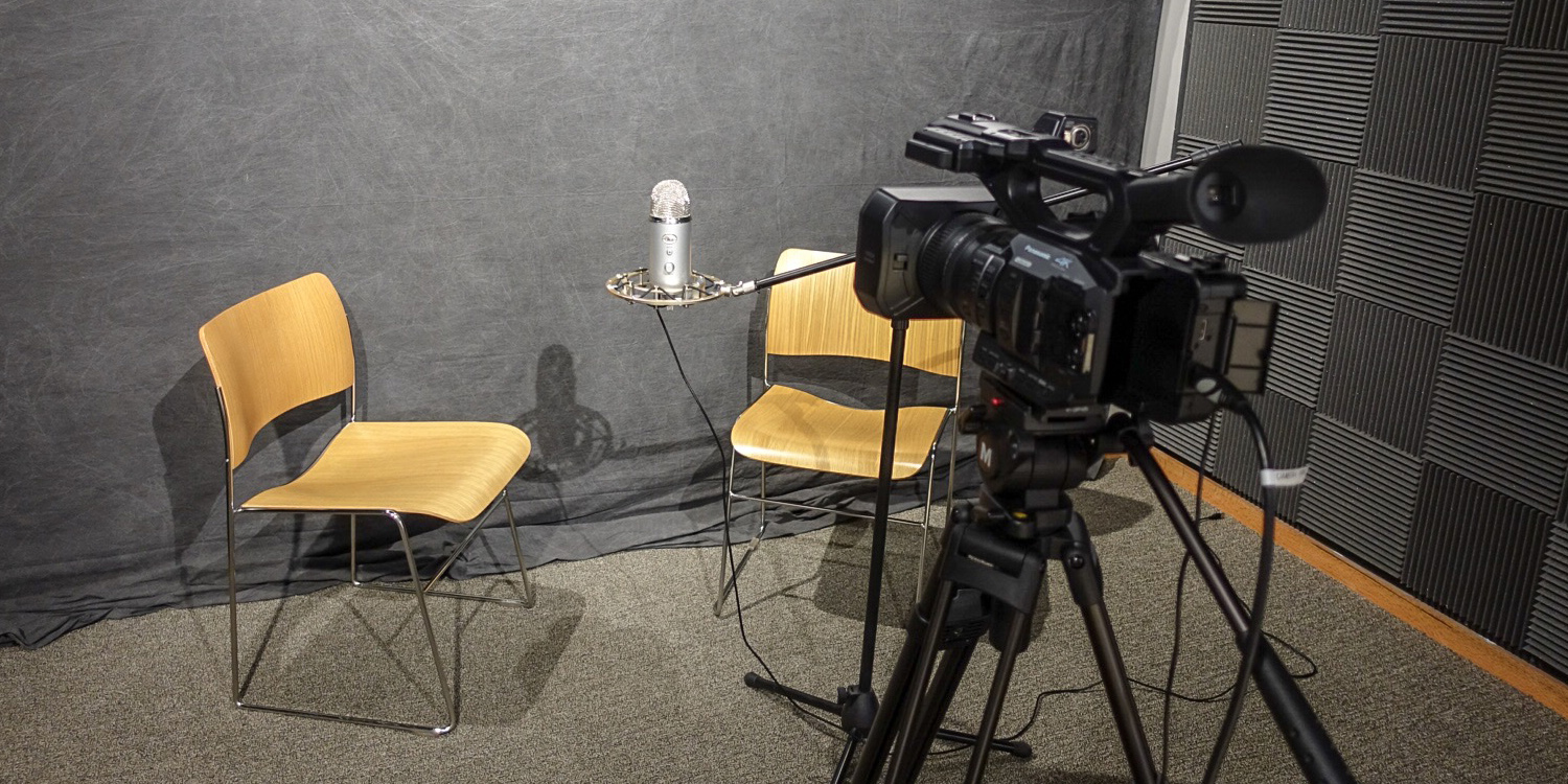 Two chairs, a microphone, and a camera are in the middle of the recording studio.
